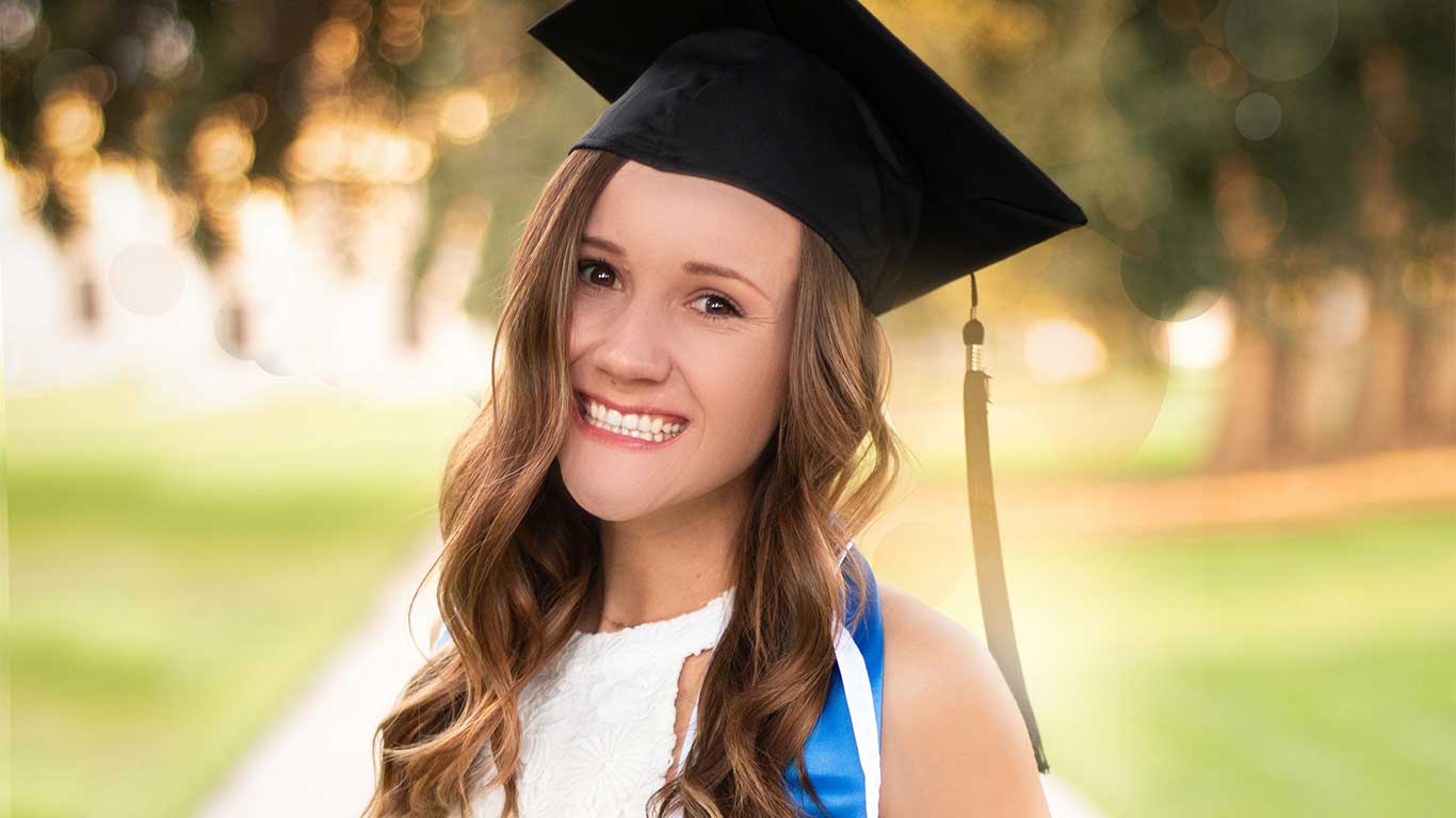 A young woman outside smiling at the camera wearing her high school graduation cap and sash.